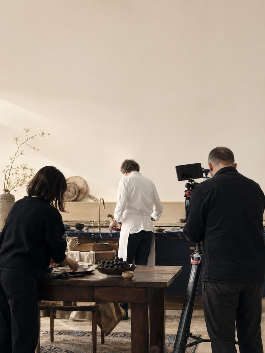 A four-person film crew lighting a tabletop set in the Valcourt studio, the camera operator framing a shot across a long walnut table.