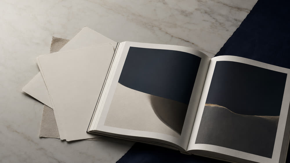An open magazine spread and loose proof sheets laid on a marble table in raking afternoon light.