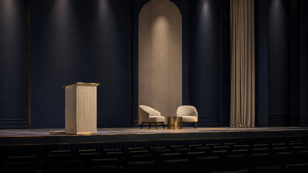 A brass lectern on a low stage in a darkened summit hall, rows of empty seats waiting in front of it.
